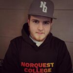 A young man with a brown beard is wearing a black baseball cap with the letters NQ on it, and a black t-shirt that says "NorQuest College" in orange and red text.