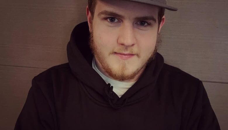 A young man with a brown beard is wearing a black baseball cap with the letters NQ on it, and a black t-shirt that says "NorQuest College" in orange and red text.